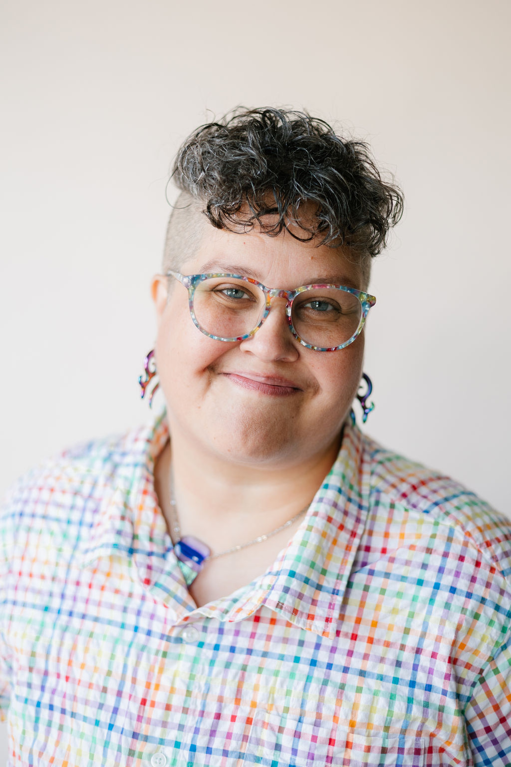 A light-brown skinned individual with brown and grey hair. They are smiling, and wearing glasses, earrings, and a button down top with white and multicolored rainbow gingham pattern.